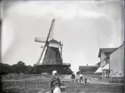 Foto av en väderkvarn med ett bostadshus till höger. I förgrunden syns en gammal kvinna och några barn.