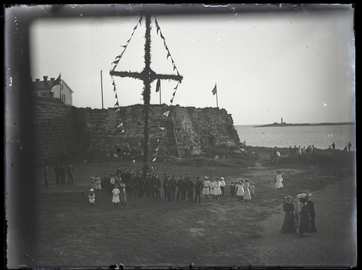 Ett kompani beväringar och några flickor har samlats vid midsommarstången (?) nedanför Varbergs fästning. Kvällsbild.
