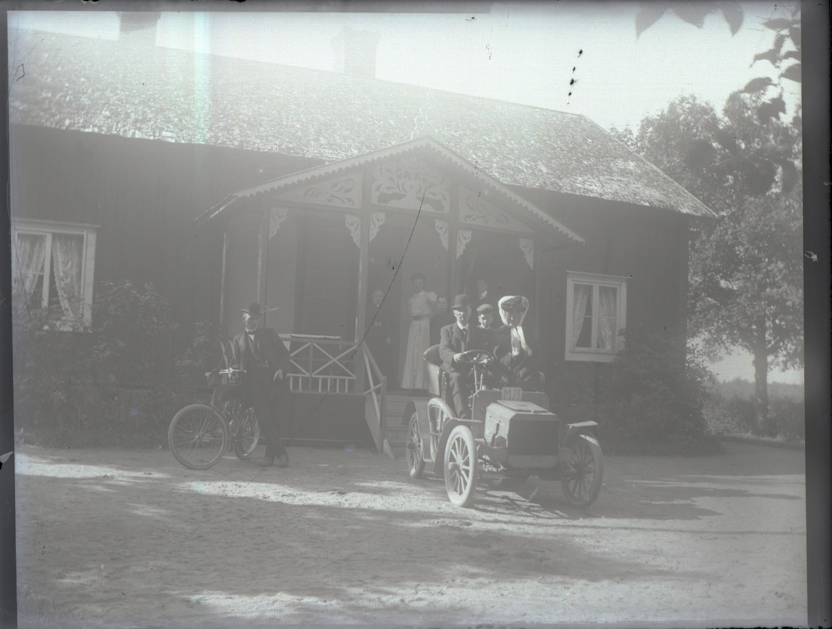 En bil med registreringsnummer M 79 är parkerad framför ett bostadshus på landet, troligen i närheten av Urshult.
Bilen är en Scania från cirka 1903, som ägdes av en Johan Sjögren. Bilen fick senare reg.nr. G 5 och skrotades 1919.
Vid cykeln står Johans son Gustaf Sjögren.