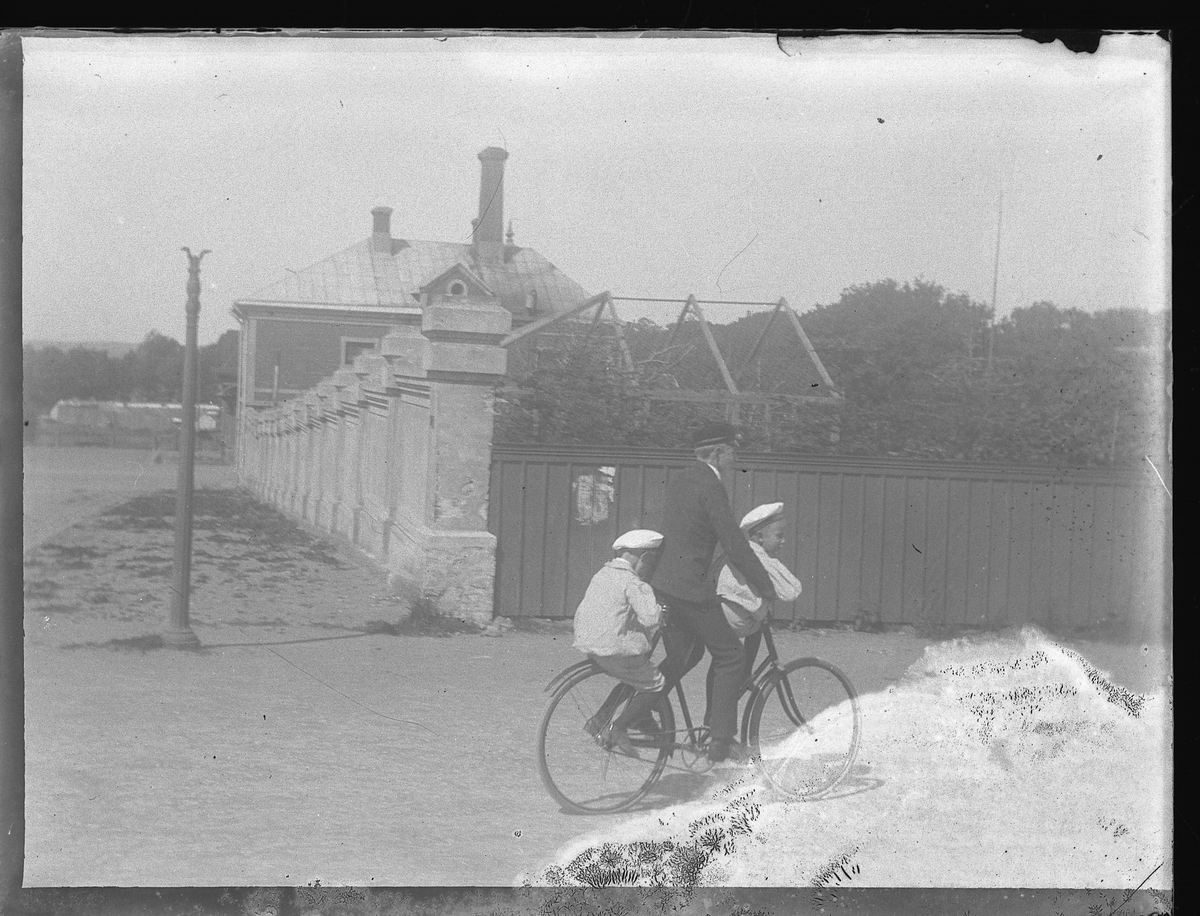 Foto av en tonårspojke, som skjutsar två pojkar på en cykel. 
I bakgrunden ser man en trädgård, omgärdad av en mur och ett plank.