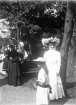 Foto av en ung kvinna i stor hatt och ljus klänning.
Bredvid henne syns en flicka i ljus sommarklänning och stråhatt. 
I bakgrunden syns en tombola och några andra kvinnor, varav en med parasoll. 
Fotot trol. taget  i Societetsparken, i samband med Barnens Dag el. likn.