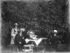 Fyra personer som samlats kring ett kaffebord utomhus.
(En av damerna syns knappt under lövverket). 
Fotografen har av allt att döma lämnat kvar sin panamahatt på kaffebordet.