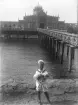 En pojke i sjömanskostym, som håller en hundvalp i famnen. 
Han står på stranden nedanför bryggan ut till kallbadhuset i Varberg.
