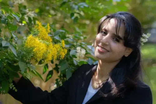 Portrett av en smilende kvinne med mørkt hår som står inntil en blomstrende busk.