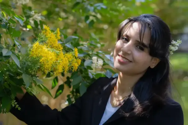 Portrett av en smilende kvinne med mørkt hår som står inntil en blomstrende busk.