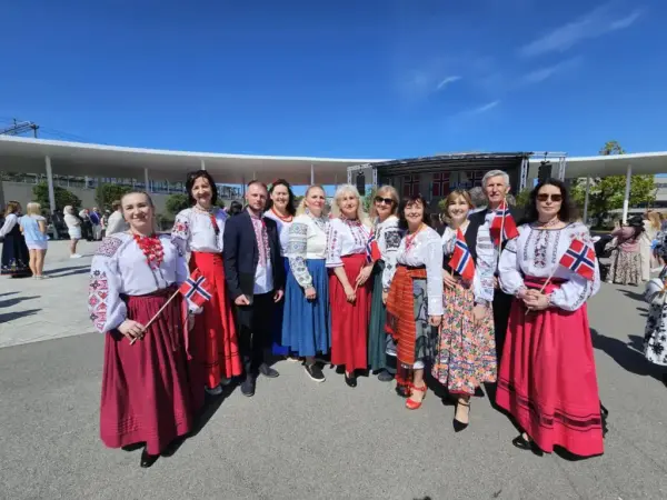 En gruppe kvinner står utendørs i fargerike kostymer. Noen holder norske flagg.