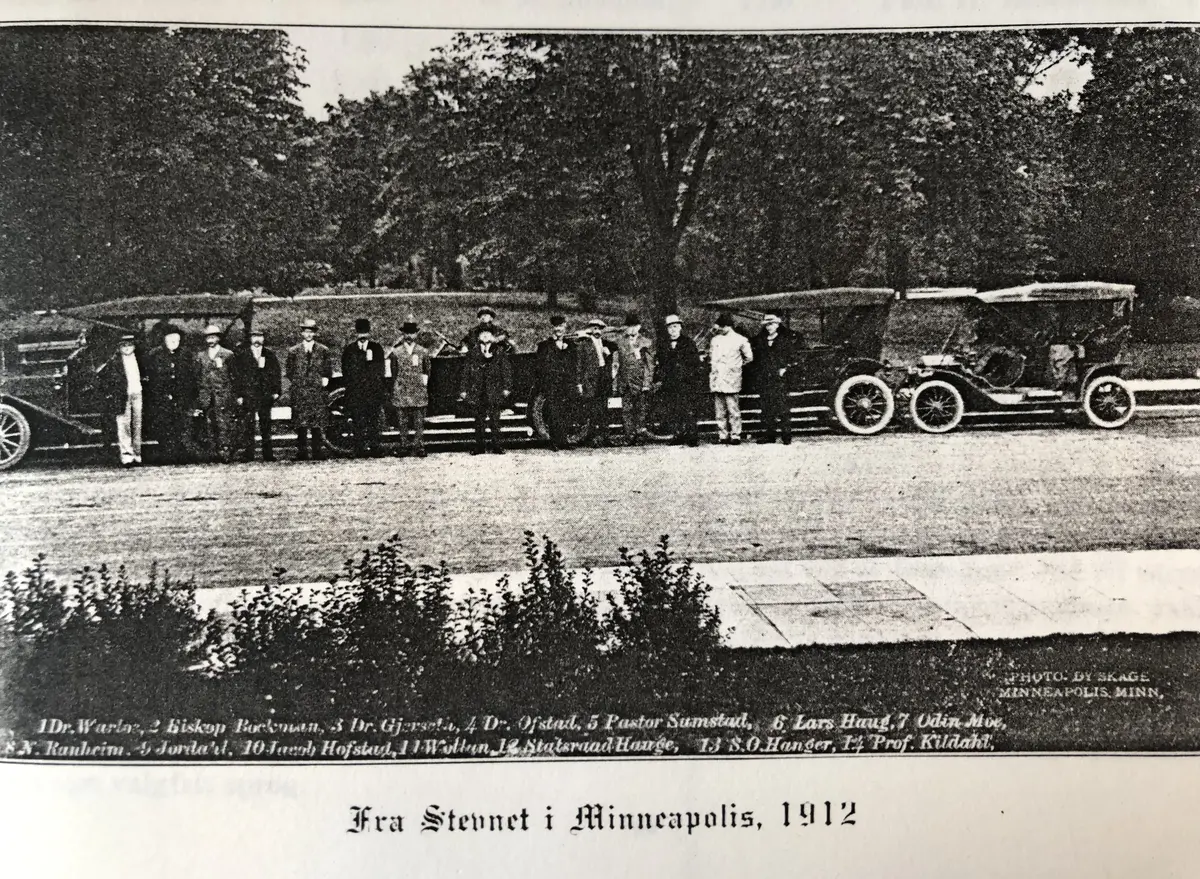 Historisk bilde av en rekke menn med hatt som står foran fire biler.