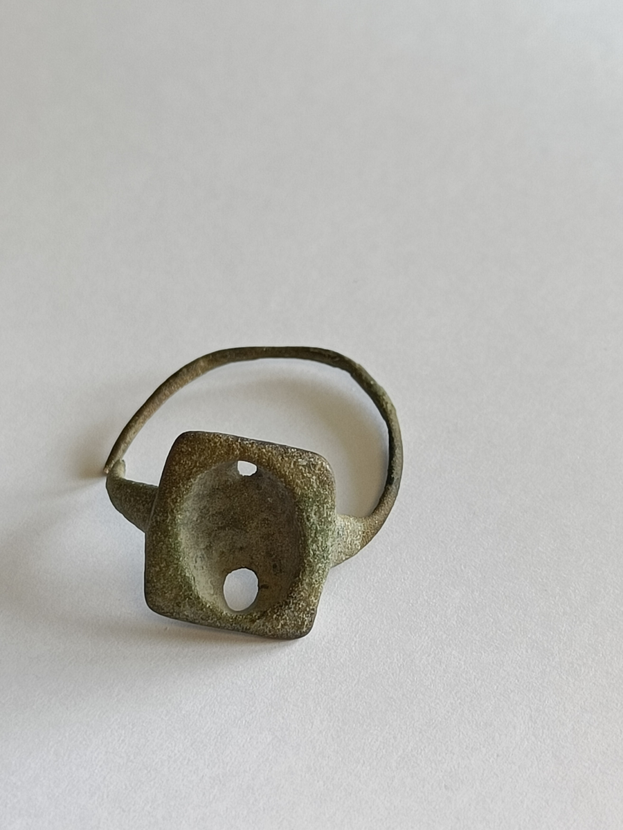 Fingerring laget av en nokså smal metalltråd med rundt tverrsnitt og et bruddsted. Gjenstanden har en rektangulær plate med en oval fordypning som trolig har vært fattet med en stein, et glass eller noe liknenede. I platen sees to hull, minst ett av dem er trolig til feste av det som en gang var fattet i ringen.