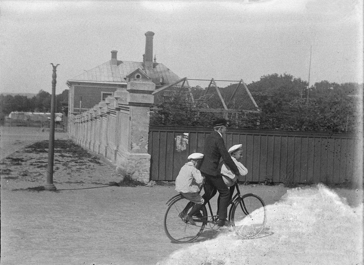 Foto av en tonårspojke, som skjutsar två pojkar på en cykel. 
I bakgrunden ser man en trädgård, omgärdad av en mur och ett plank.
