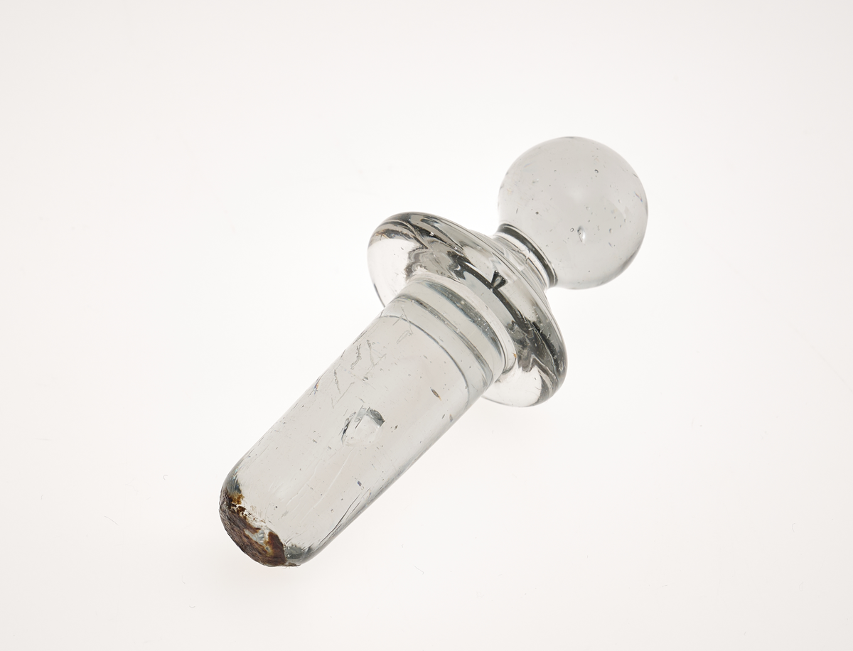 En glasspropp som trolig har tilhørt en karaffel. Den er laget av klart glass med hint ab gråfarge i. Øverst er det en kule. Deretter er det en mansjett.Til sist er korkdelen som er sylindrisk på form (men ikke hul). I proppen kan man se luftbobler etter produksjon.
