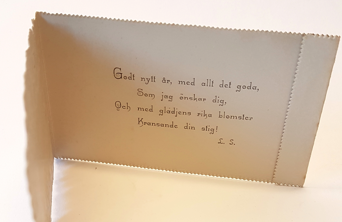 Nyårskort med motiv av violer i lila och blå toner och detaljer i guld. Godt Nytt år i guldskrift i kortets mitt.  På insidan en tryckt nyårshälsning, signerad L.S