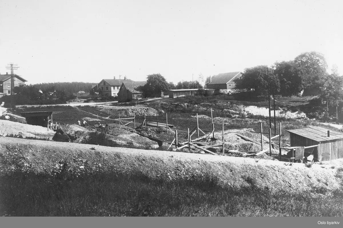 Bildet tatt i forbindelse med utbyggingen av Lilleakerbanen som ble åpnet i 1919, villabebyggelse og gårdsbruk i bakgrunnen. Gårdsbruket til høyre i bakgrunnen er Ørakerbråten, også kalt Schlægerbråten etter glassmester Schlæger som bodde der. Gården ligger der fortsatt like ved nåværende Lilleaker trikkestopp. Til venstre i bildet sees en av arbeiderboligene - Sagførergården, gnr 10, br.nr. 35.