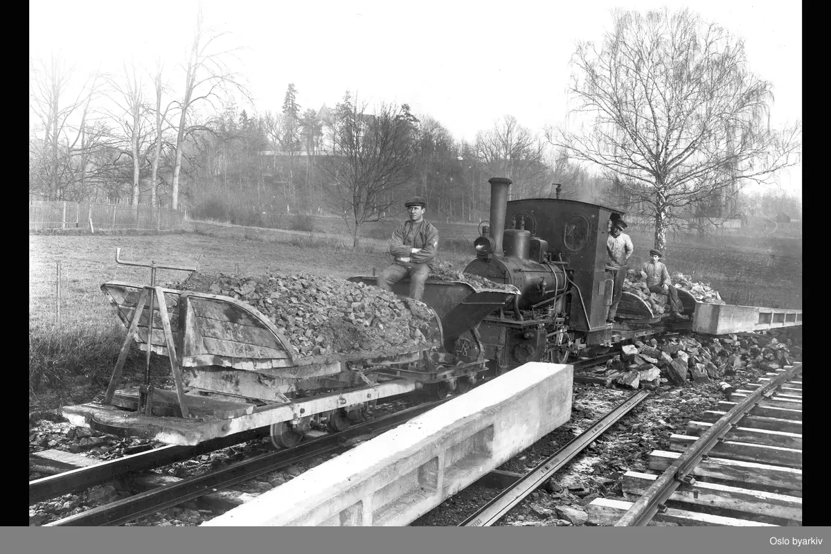 Bildet er tatt under utbyggingen av Lilleakerbanen. Kjøring av grus, stein, betongblokker ligger på skinnene, arbeidere, dyrket mark i bakgrunnen.