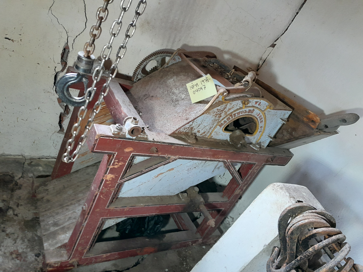 Piggtreskemaskin. Kontruksjon (stativ) i tre rundt maskina. 
Øvste del av maskina er i metall, medan nedste (renne) er i tre. Maskina har opning oppe på eine kortsida, der det er monternt ein metall sylinder med piggar (på aksling). Under metall sylindaren er det ei trerenne som skrånar nedover. 
På eine sida av stativet er det montert fleire tannhjul som grip i kvarandre. Stort hjul i tre ytst. Feste for sveiv stikk ut på andre sida. 
Sveiva driv hjulet via aksling under tre-renna, og via tannhjule vil den pigga metallsylinderen dreie rundt.
Treråma er raudlilla med kvit og svart stripe. Maskina er turkis med gule kantar og raud skrift på sidene.