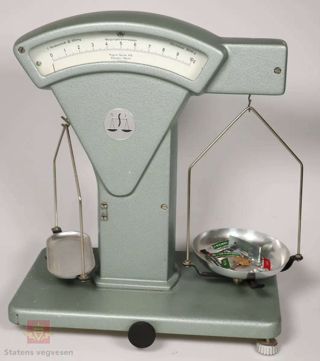 Grønn vekt som er laget hovedsakelig av metall. Vekten har to skåler på hver side som kan hektes av og på. I en av skålene ligger det skruer, et par nøkler og flere merkelapper som mest sannsynlig kan ha hørt til gjenstanden.