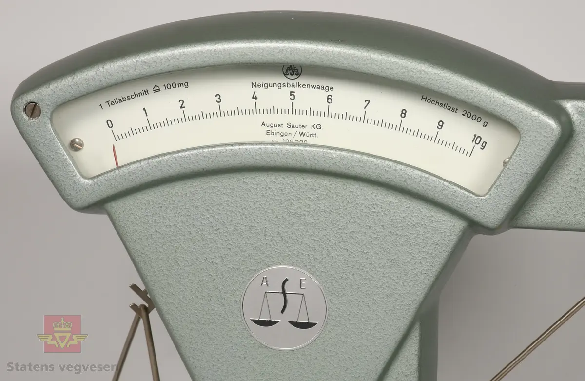 Grønn vekt som er laget hovedsakelig av metall. Vekten har to skåler på hver side som kan hektes av og på. I en av skålene ligger det skruer, et par nøkler og flere merkelapper som mest sannsynlig kan ha hørt til gjenstanden.