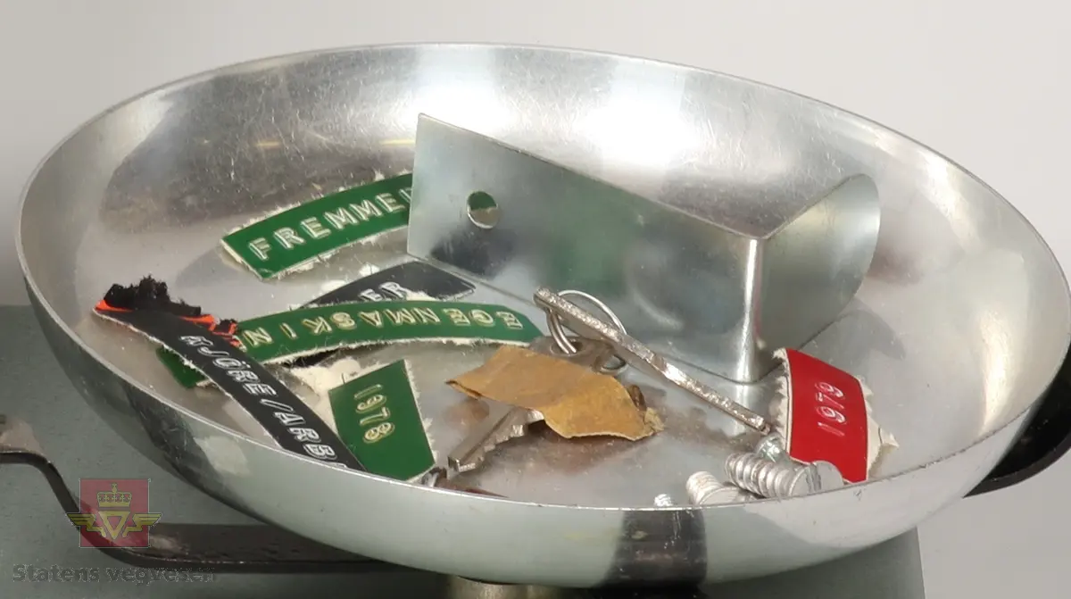 Grønn vekt som er laget hovedsakelig av metall. Vekten har to skåler på hver side som kan hektes av og på. I en av skålene ligger det skruer, et par nøkler og flere merkelapper som mest sannsynlig kan ha hørt til gjenstanden.