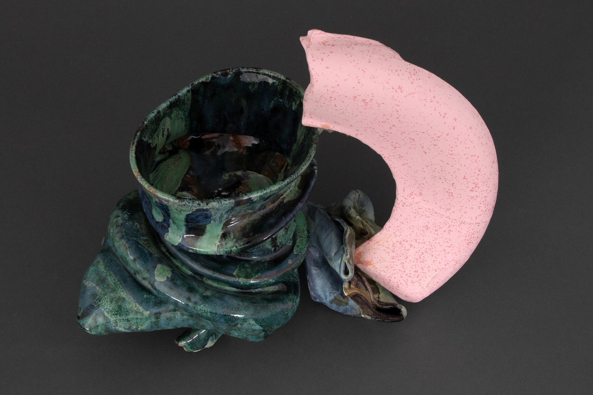 Dreid leirgodskropp som er deformert i etterkant. Reultatet er at den tukturelle stramheten i korpus bryter sammen og delvis folder seg. Spettet grønn og blå glasur med innsalg av brunt på korpus. Rosa porslenshank. Formen har referanser til kannen.