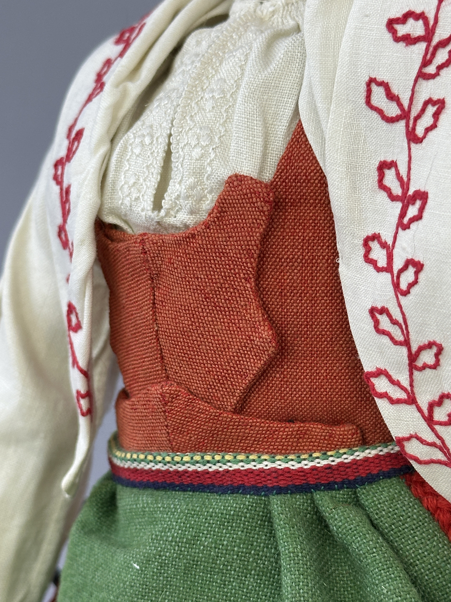 Docka klädd i kvinnlig sockendräkt från Norra Ny i Värmland. Kroppen är sydd i linnetyg och stoppad med textilt material. Huvud och händer är av lergods. Kläder sydda av handvävda tyger förutom skjorta, underkjol och halskläde, som är maskinvävt. Halskläde är broderat med tambursöm, skjortan med plattsöm och myrgång samt har stripade veck runt halskrage och manschetter. Även kjolen har lagda veck runtom och är fastsytt med livstycket. Förklädet, med 3 lagda veck på var sida, är kantad runtom med en tre-fläta gjord av rött ullgarn, vilket också kjolen har i nederkanten. Ullstrumporna är handstickade i slätstickning med en kant upptill i 2-2 resår och broderad med förstygn av rött ullgarn. Huvudet är prytt med hår av lintågor, som delvis är flätat till hårflätor samt ett målat hårnäver. Dockans fötter bär svarta skinnskor.