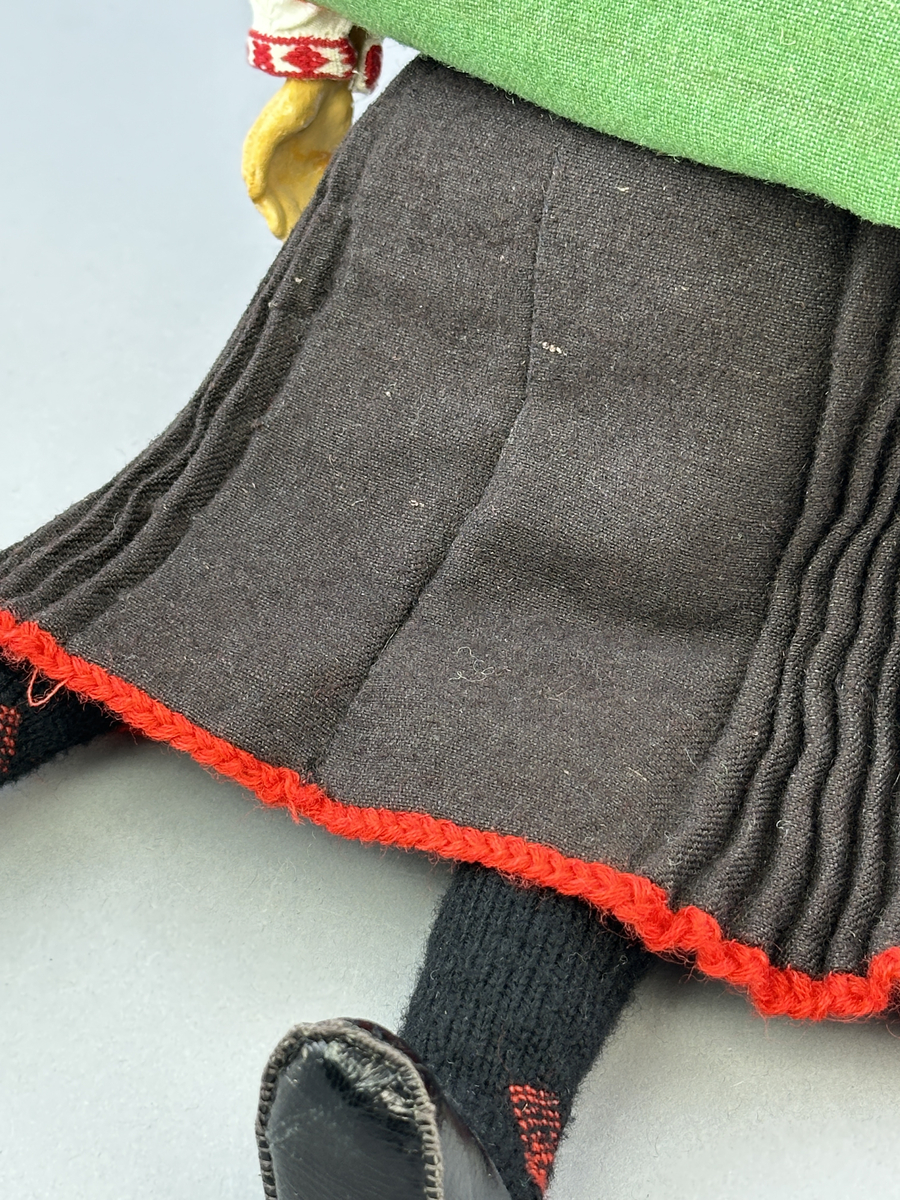 Docka klädd i kvinnlig sockendräkt från Norra Ny i Värmland. Kroppen är sydd i linnetyg och stoppad med textilt material. Huvud och händer är av lergods. Kläder sydda av handvävda tyger förutom skjorta, underkjol och halskläde, som är maskinvävt. Halskläde är broderat med tambursöm, skjortan med plattsöm och myrgång samt har stripade veck runt halskrage och manschetter. Även kjolen har lagda veck runtom och är fastsytt med livstycket. Förklädet, med 3 lagda veck på var sida, är kantad runtom med en tre-fläta gjord av rött ullgarn, vilket också kjolen har i nederkanten. Ullstrumporna är handstickade i slätstickning med en kant upptill i 2-2 resår och broderad med förstygn av rött ullgarn. Huvudet är prytt med hår av lintågor, som delvis är flätat till hårflätor samt ett målat hårnäver. Dockans fötter bär svarta skinnskor.