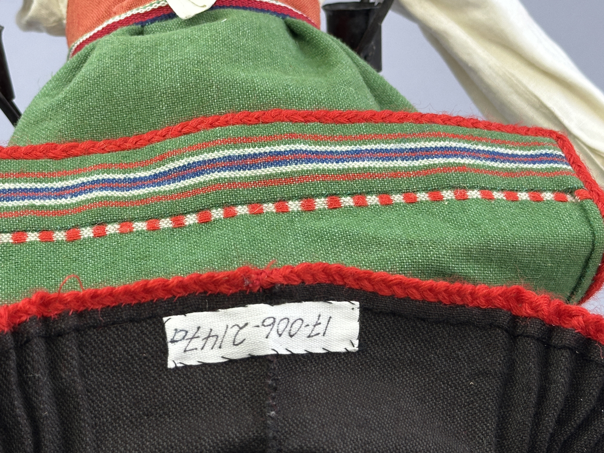 Docka klädd i kvinnlig sockendräkt från Norra Ny i Värmland. Kroppen är sydd i linnetyg och stoppad med textilt material. Huvud och händer är av lergods. Kläder sydda av handvävda tyger förutom skjorta, underkjol och halskläde, som är maskinvävt. Halskläde är broderat med tambursöm, skjortan med plattsöm och myrgång samt har stripade veck runt halskrage och manschetter. Även kjolen har lagda veck runtom och är fastsytt med livstycket. Förklädet, med 3 lagda veck på var sida, är kantad runtom med en tre-fläta gjord av rött ullgarn, vilket också kjolen har i nederkanten. Ullstrumporna är handstickade i slätstickning med en kant upptill i 2-2 resår och broderad med förstygn av rött ullgarn. Huvudet är prytt med hår av lintågor, som delvis är flätat till hårflätor samt ett målat hårnäver. Dockans fötter bär svarta skinnskor.