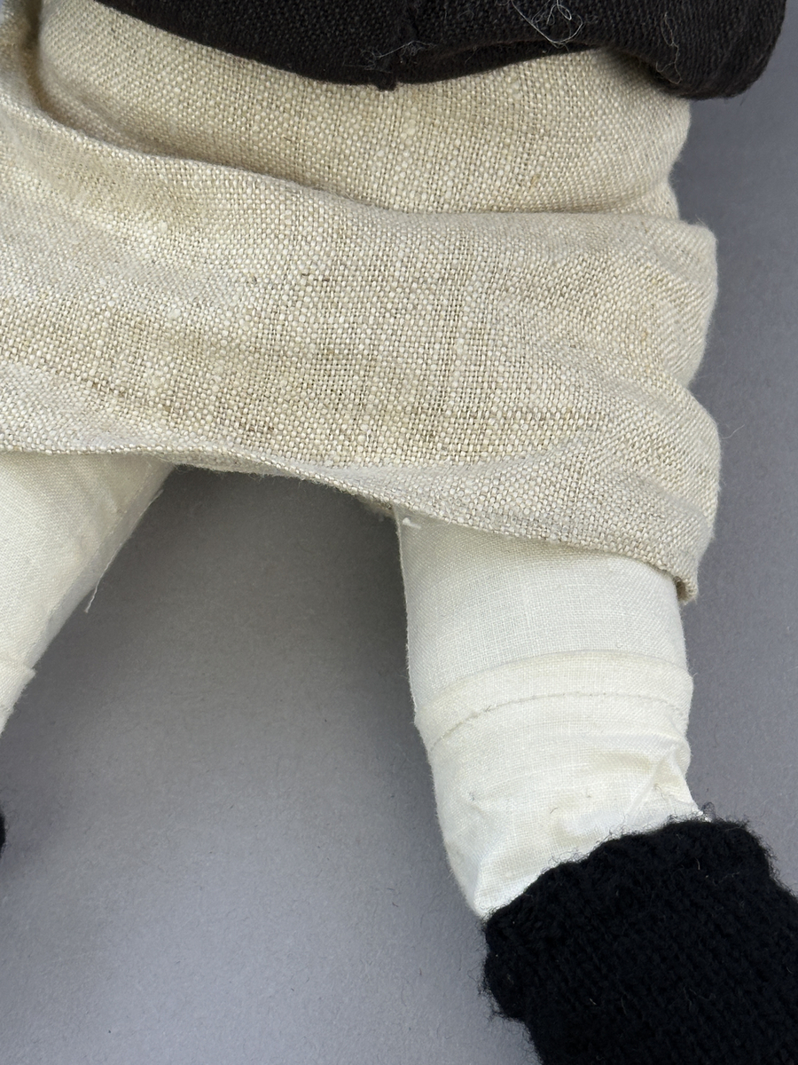 Docka klädd i kvinnlig sockendräkt från Norra Ny i Värmland. Kroppen är sydd i linnetyg och stoppad med textilt material. Huvud och händer är av lergods. Kläder sydda av handvävda tyger förutom skjorta, underkjol och halskläde, som är maskinvävt. Halskläde är broderat med tambursöm, skjortan med plattsöm och myrgång samt har stripade veck runt halskrage och manschetter. Även kjolen har lagda veck runtom och är fastsytt med livstycket. Förklädet, med 3 lagda veck på var sida, är kantad runtom med en tre-fläta gjord av rött ullgarn, vilket också kjolen har i nederkanten. Ullstrumporna är handstickade i slätstickning med en kant upptill i 2-2 resår och broderad med förstygn av rött ullgarn. Huvudet är prytt med hår av lintågor, som delvis är flätat till hårflätor samt ett målat hårnäver. Dockans fötter bär svarta skinnskor.