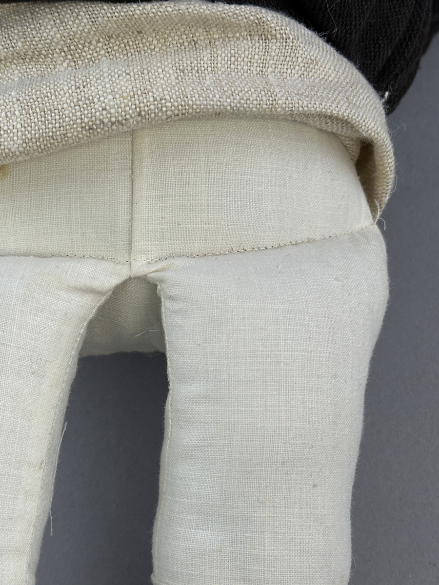 Docka klädd i kvinnlig sockendräkt från Norra Ny i Värmland. Kroppen är sydd i linnetyg och stoppad med textilt material. Huvud och händer är av lergods. Kläder sydda av handvävda tyger förutom skjorta, underkjol och halskläde, som är maskinvävt. Halskläde är broderat med tambursöm, skjortan med plattsöm och myrgång samt har stripade veck runt halskrage och manschetter. Även kjolen har lagda veck runtom och är fastsytt med livstycket. Förklädet, med 3 lagda veck på var sida, är kantad runtom med en tre-fläta gjord av rött ullgarn, vilket också kjolen har i nederkanten. Ullstrumporna är handstickade i slätstickning med en kant upptill i 2-2 resår och broderad med förstygn av rött ullgarn. Huvudet är prytt med hår av lintågor, som delvis är flätat till hårflätor samt ett målat hårnäver. Dockans fötter bär svarta skinnskor.