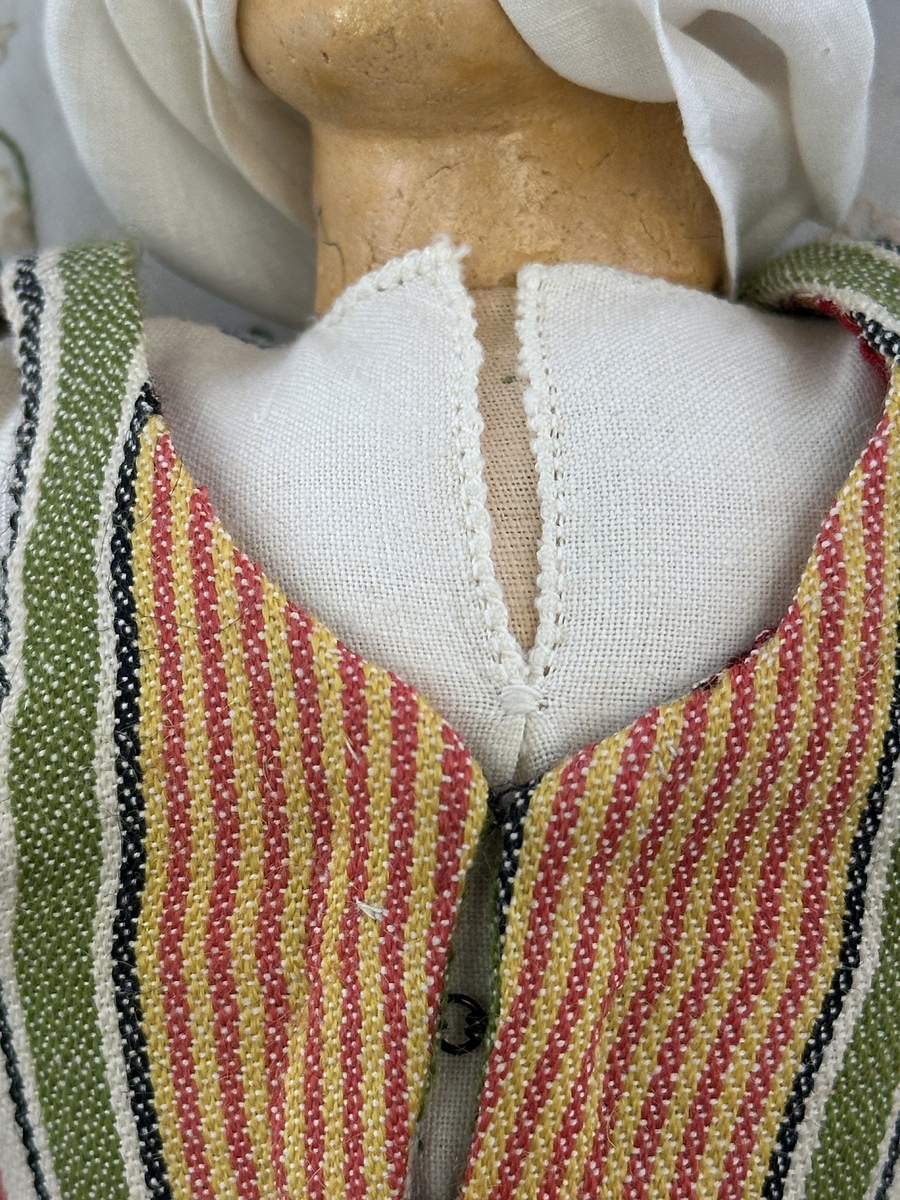 Docka klädd i kvinnlig sockendräkt från Jösse härad i Värmland. Kroppen är handsydd i linnetyg och stoppad med textilt material. Huvud och händer är av lergods. Kläder handsydda av handvävda tyger förutom skjorta, underkjol och halskläde, som är maskinvävt. Halskläde är broderat med tambursöm, skjorta med myrgång runt halskant, sprund och manschett. Skjortan har också ett blått korsstygnsbroderi på axel och manschett samt stripade veck runt  manschetter. Halsklädet hålls ihop av en sölja i nysilver med graverat mönster. Kjol sydd med lagda veck runtom, utom under förkläde, vars linning består av ett bomullsband. Kjolen är också dekorerad med fyra tre-flätor av rött ullgarn i nederkant. Förklädet, är rikt broderat med utdragssöm, plattsöm, hålsöm och langettsöm, har stripade veck vid midja samt är kantad med en handknypplad spets. Tyget till livstycket har bomullsvarp och inslag av redgarn i grönt, gult, svart, vitt samt två olika rosa toner. Livstycket är vändsytt/fodrat med ett rutigt bomullstyg och knäpps med 5 hyskar & hakar. Ullstrumporna är handstickade i slätstickning med en kant upptill i 2-2 resår och broderad med förstygn/plattsöm i rött bomullsgarn och grönt ullgarn. Huvudet är prytt med en blå bindhätta av siden kantad med ett tyllbroderi i vitt och fyra hängande ljusgröna sidenband baktill cirka 12cm långa. Dockans fötter bär svarta skinnskor.