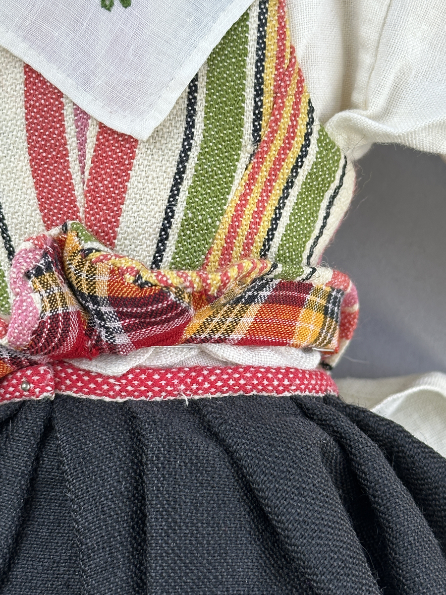 Docka klädd i kvinnlig sockendräkt från Jösse härad i Värmland. Kroppen är handsydd i linnetyg och stoppad med textilt material. Huvud och händer är av lergods. Kläder handsydda av handvävda tyger förutom skjorta, underkjol och halskläde, som är maskinvävt. Halskläde är broderat med tambursöm, skjorta med myrgång runt halskant, sprund och manschett. Skjortan har också ett blått korsstygnsbroderi på axel och manschett samt stripade veck runt  manschetter. Halsklädet hålls ihop av en sölja i nysilver med graverat mönster. Kjol sydd med lagda veck runtom, utom under förkläde, vars linning består av ett bomullsband. Kjolen är också dekorerad med fyra tre-flätor av rött ullgarn i nederkant. Förklädet, är rikt broderat med utdragssöm, plattsöm, hålsöm och langettsöm, har stripade veck vid midja samt är kantad med en handknypplad spets. Tyget till livstycket har bomullsvarp och inslag av redgarn i grönt, gult, svart, vitt samt två olika rosa toner. Livstycket är vändsytt/fodrat med ett rutigt bomullstyg och knäpps med 5 hyskar & hakar. Ullstrumporna är handstickade i slätstickning med en kant upptill i 2-2 resår och broderad med förstygn/plattsöm i rött bomullsgarn och grönt ullgarn. Huvudet är prytt med en blå bindhätta av siden kantad med ett tyllbroderi i vitt och fyra hängande ljusgröna sidenband baktill cirka 12cm långa. Dockans fötter bär svarta skinnskor.