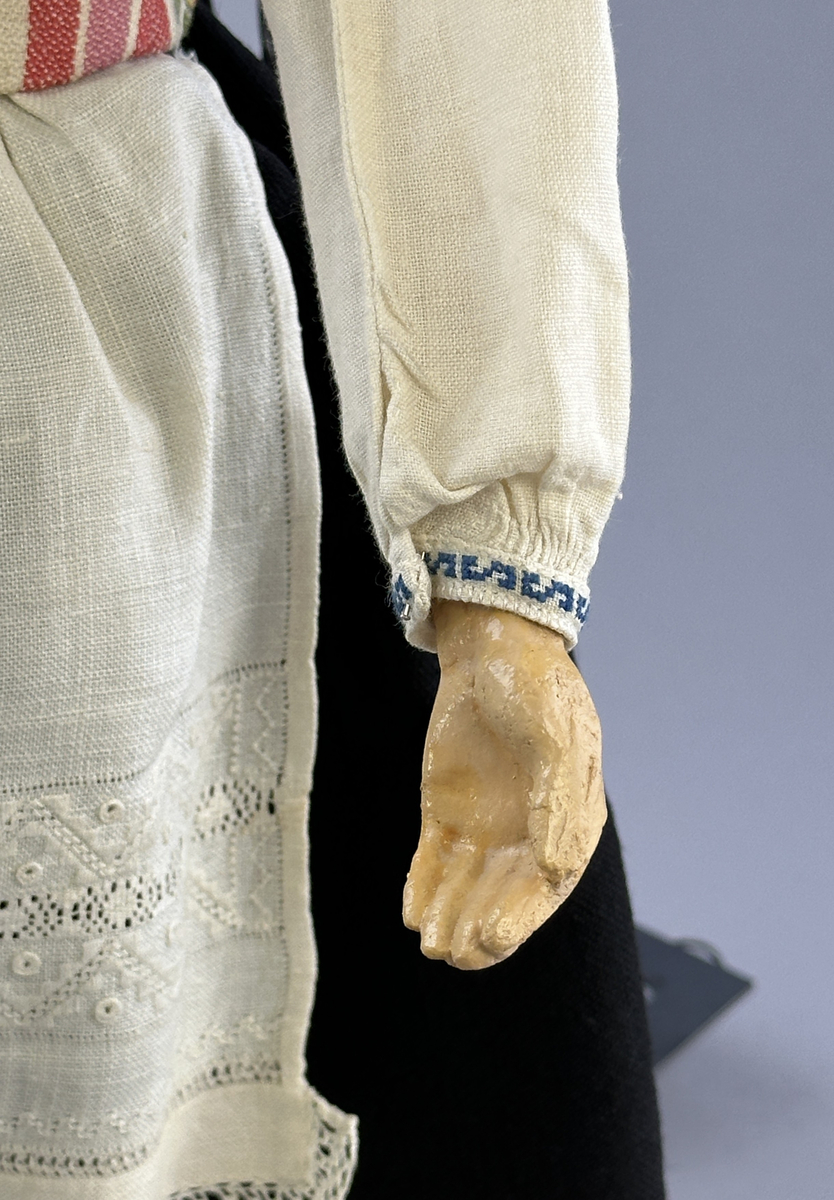 Docka klädd i kvinnlig sockendräkt från Jösse härad i Värmland. Kroppen är handsydd i linnetyg och stoppad med textilt material. Huvud och händer är av lergods. Kläder handsydda av handvävda tyger förutom skjorta, underkjol och halskläde, som är maskinvävt. Halskläde är broderat med tambursöm, skjorta med myrgång runt halskant, sprund och manschett. Skjortan har också ett blått korsstygnsbroderi på axel och manschett samt stripade veck runt  manschetter. Halsklädet hålls ihop av en sölja i nysilver med graverat mönster. Kjol sydd med lagda veck runtom, utom under förkläde, vars linning består av ett bomullsband. Kjolen är också dekorerad med fyra tre-flätor av rött ullgarn i nederkant. Förklädet, är rikt broderat med utdragssöm, plattsöm, hålsöm och langettsöm, har stripade veck vid midja samt är kantad med en handknypplad spets. Tyget till livstycket har bomullsvarp och inslag av redgarn i grönt, gult, svart, vitt samt två olika rosa toner. Livstycket är vändsytt/fodrat med ett rutigt bomullstyg och knäpps med 5 hyskar & hakar. Ullstrumporna är handstickade i slätstickning med en kant upptill i 2-2 resår och broderad med förstygn/plattsöm i rött bomullsgarn och grönt ullgarn. Huvudet är prytt med en blå bindhätta av siden kantad med ett tyllbroderi i vitt och fyra hängande ljusgröna sidenband baktill cirka 12cm långa. Dockans fötter bär svarta skinnskor.