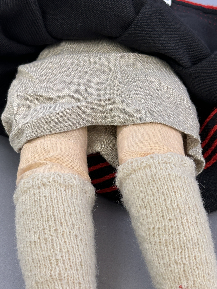 Docka klädd i kvinnlig sockendräkt från Jösse härad i Värmland. Kroppen är handsydd i linnetyg och stoppad med textilt material. Huvud och händer är av lergods. Kläder handsydda av handvävda tyger förutom skjorta, underkjol och halskläde, som är maskinvävt. Halskläde är broderat med tambursöm, skjorta med myrgång runt halskant, sprund och manschett. Skjortan har också ett blått korsstygnsbroderi på axel och manschett samt stripade veck runt  manschetter. Halsklädet hålls ihop av en sölja i nysilver med graverat mönster. Kjol sydd med lagda veck runtom, utom under förkläde, vars linning består av ett bomullsband. Kjolen är också dekorerad med fyra tre-flätor av rött ullgarn i nederkant. Förklädet, är rikt broderat med utdragssöm, plattsöm, hålsöm och langettsöm, har stripade veck vid midja samt är kantad med en handknypplad spets. Tyget till livstycket har bomullsvarp och inslag av redgarn i grönt, gult, svart, vitt samt två olika rosa toner. Livstycket är vändsytt/fodrat med ett rutigt bomullstyg och knäpps med 5 hyskar & hakar. Ullstrumporna är handstickade i slätstickning med en kant upptill i 2-2 resår och broderad med förstygn/plattsöm i rött bomullsgarn och grönt ullgarn. Huvudet är prytt med en blå bindhätta av siden kantad med ett tyllbroderi i vitt och fyra hängande ljusgröna sidenband baktill cirka 12cm långa. Dockans fötter bär svarta skinnskor.