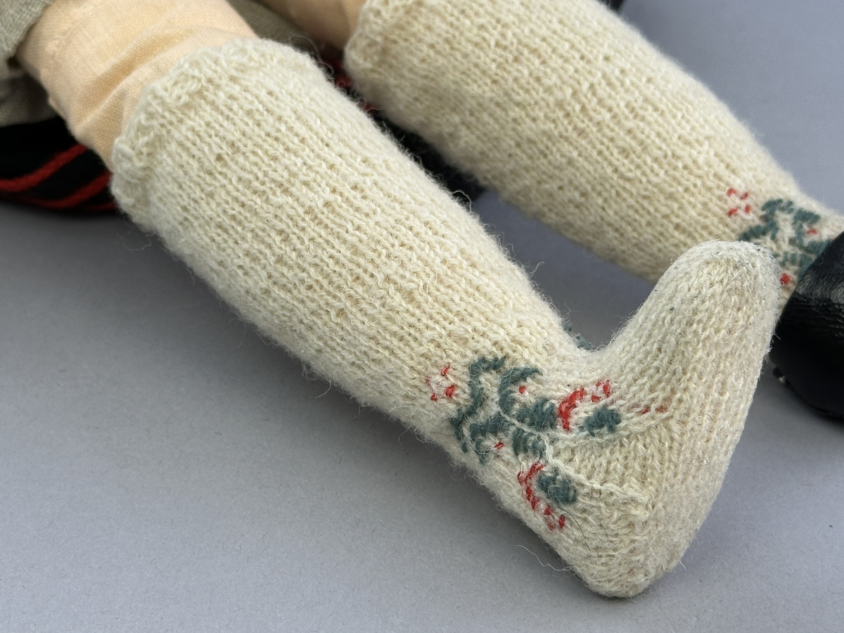 Docka klädd i kvinnlig sockendräkt från Jösse härad i Värmland. Kroppen är handsydd i linnetyg och stoppad med textilt material. Huvud och händer är av lergods. Kläder handsydda av handvävda tyger förutom skjorta, underkjol och halskläde, som är maskinvävt. Halskläde är broderat med tambursöm, skjorta med myrgång runt halskant, sprund och manschett. Skjortan har också ett blått korsstygnsbroderi på axel och manschett samt stripade veck runt  manschetter. Halsklädet hålls ihop av en sölja i nysilver med graverat mönster. Kjol sydd med lagda veck runtom, utom under förkläde, vars linning består av ett bomullsband. Kjolen är också dekorerad med fyra tre-flätor av rött ullgarn i nederkant. Förklädet, är rikt broderat med utdragssöm, plattsöm, hålsöm och langettsöm, har stripade veck vid midja samt är kantad med en handknypplad spets. Tyget till livstycket har bomullsvarp och inslag av redgarn i grönt, gult, svart, vitt samt två olika rosa toner. Livstycket är vändsytt/fodrat med ett rutigt bomullstyg och knäpps med 5 hyskar & hakar. Ullstrumporna är handstickade i slätstickning med en kant upptill i 2-2 resår och broderad med förstygn/plattsöm i rött bomullsgarn och grönt ullgarn. Huvudet är prytt med en blå bindhätta av siden kantad med ett tyllbroderi i vitt och fyra hängande ljusgröna sidenband baktill cirka 12cm långa. Dockans fötter bär svarta skinnskor.