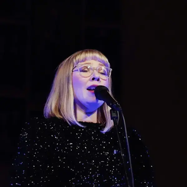 Portrett av en kvinne med briller og lyst, halvlangt  hår som synger i en mikrofon. Mørk kjole og mørk bakgrunn.