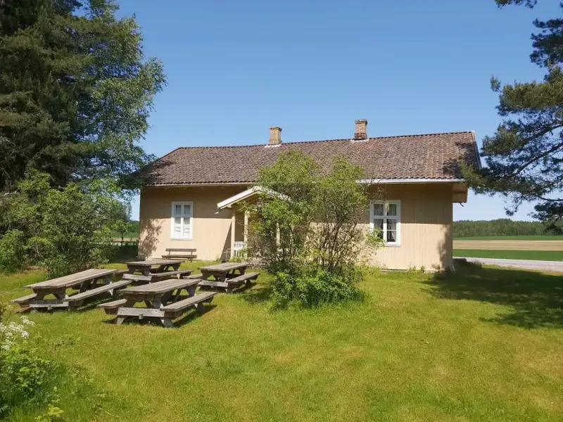 Mindre skolehus fra 1860-årene med hage foran. Fire piknikbenker. Blå himmel.