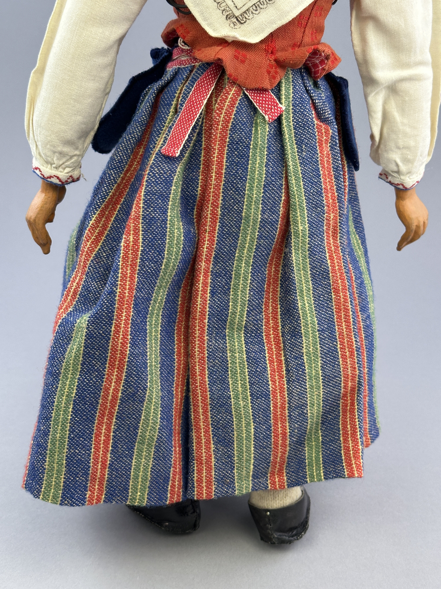 Docka klädd i kvinnlig sockendräkt från Östervallskogs socken i Värmland. Kroppen är handsydd i linnetyg och stoppad med textilt material. Huvud och händer är av lergods. Kläder handsydda av handvävda tyger förutom skjorta, underkjol och halskläde, som är maskinvävt. Skjorta är broderad med myrgång runt halssprund, plattsöm på hals- och handlinning samt en blå stjälksöm i kanten på handlinningen. Halsklädet, med tryck i gult, rosa och beige, hålls ihop av en sölja i stål med punsat mönster. Tyget till kjolen har oblekt linvarp med ullinslag i blått, grönt, rött och oblekt. Kjol sydd med lagda veck runtom, utom under förkläde, vars linning består av ett bomullsband. Förkläde, vävt med bomullsvarp och ullinslag i rött och vitt, har också lagda veck vid midja och bomullsband i rips till knytband. I midjan sitter en väska i blå och röd vadmal. Livstycket, vävt med bomullsvarp och lininslag, är vändsytt/fodrat med ett rutigt bomullstyg och knäpps med 5 hyskar & hakar. Ullstrumporna är handstickade i slätstickning med en kant upptill i 2-2 resår. Huvudet är prytt med en vit linnehätta kantad med en knypplad linnespets. Dockans fötter bär svarta skinnskor.

