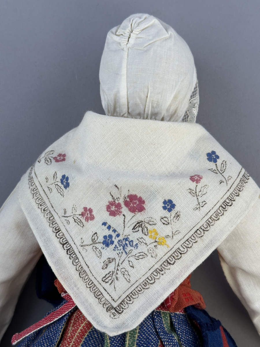 Docka klädd i kvinnlig sockendräkt från Östervallskogs socken i Värmland. Kroppen är handsydd i linnetyg och stoppad med textilt material. Huvud och händer är av lergods. Kläder handsydda av handvävda tyger förutom skjorta, underkjol och halskläde, som är maskinvävt. Skjorta är broderad med myrgång runt halssprund, plattsöm på hals- och handlinning samt en blå stjälksöm i kanten på handlinningen. Halsklädet, med tryck i gult, rosa och beige, hålls ihop av en sölja i stål med punsat mönster. Tyget till kjolen har oblekt linvarp med ullinslag i blått, grönt, rött och oblekt. Kjol sydd med lagda veck runtom, utom under förkläde, vars linning består av ett bomullsband. Förkläde, vävt med bomullsvarp och ullinslag i rött och vitt, har också lagda veck vid midja och bomullsband i rips till knytband. I midjan sitter en väska i blå och röd vadmal. Livstycket, vävt med bomullsvarp och lininslag, är vändsytt/fodrat med ett rutigt bomullstyg och knäpps med 5 hyskar & hakar. Ullstrumporna är handstickade i slätstickning med en kant upptill i 2-2 resår. Huvudet är prytt med en vit linnehätta kantad med en knypplad linnespets. Dockans fötter bär svarta skinnskor.

