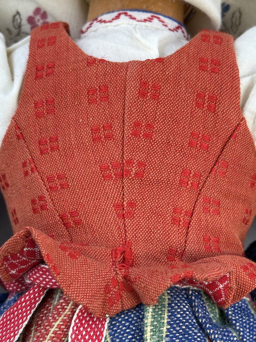 Docka klädd i kvinnlig sockendräkt från Östervallskogs socken i Värmland. Kroppen är handsydd i linnetyg och stoppad med textilt material. Huvud och händer är av lergods. Kläder handsydda av handvävda tyger förutom skjorta, underkjol och halskläde, som är maskinvävt. Skjorta är broderad med myrgång runt halssprund, plattsöm på hals- och handlinning samt en blå stjälksöm i kanten på handlinningen. Halsklädet, med tryck i gult, rosa och beige, hålls ihop av en sölja i stål med punsat mönster. Tyget till kjolen har oblekt linvarp med ullinslag i blått, grönt, rött och oblekt. Kjol sydd med lagda veck runtom, utom under förkläde, vars linning består av ett bomullsband. Förkläde, vävt med bomullsvarp och ullinslag i rött och vitt, har också lagda veck vid midja och bomullsband i rips till knytband. I midjan sitter en väska i blå och röd vadmal. Livstycket, vävt med bomullsvarp och lininslag, är vändsytt/fodrat med ett rutigt bomullstyg och knäpps med 5 hyskar & hakar. Ullstrumporna är handstickade i slätstickning med en kant upptill i 2-2 resår. Huvudet är prytt med en vit linnehätta kantad med en knypplad linnespets. Dockans fötter bär svarta skinnskor.


