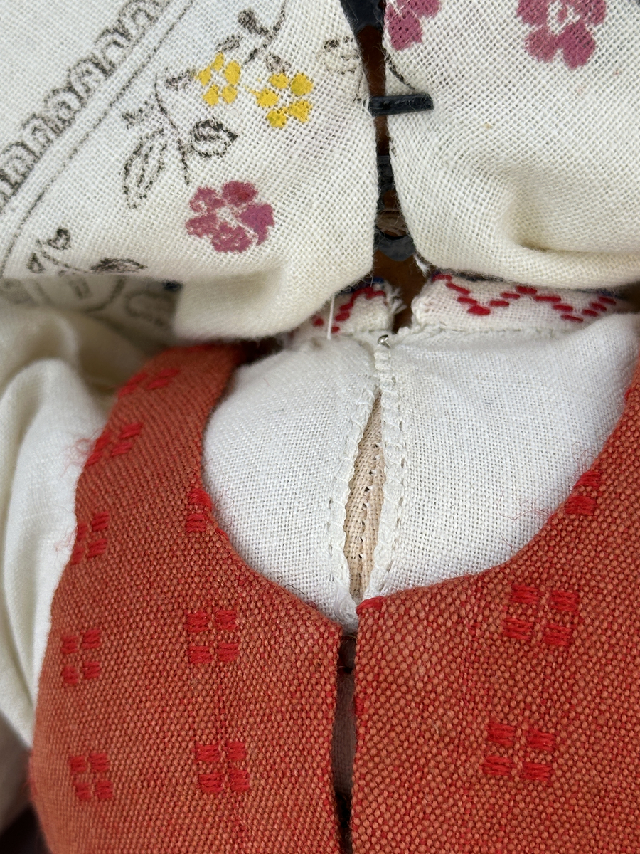 Docka klädd i kvinnlig sockendräkt från Östervallskogs socken i Värmland. Kroppen är handsydd i linnetyg och stoppad med textilt material. Huvud och händer är av lergods. Kläder handsydda av handvävda tyger förutom skjorta, underkjol och halskläde, som är maskinvävt. Skjorta är broderad med myrgång runt halssprund, plattsöm på hals- och handlinning samt en blå stjälksöm i kanten på handlinningen. Halsklädet, med tryck i gult, rosa och beige, hålls ihop av en sölja i stål med punsat mönster. Tyget till kjolen har oblekt linvarp med ullinslag i blått, grönt, rött och oblekt. Kjol sydd med lagda veck runtom, utom under förkläde, vars linning består av ett bomullsband. Förkläde, vävt med bomullsvarp och ullinslag i rött och vitt, har också lagda veck vid midja och bomullsband i rips till knytband. I midjan sitter en väska i blå och röd vadmal. Livstycket, vävt med bomullsvarp och lininslag, är vändsytt/fodrat med ett rutigt bomullstyg och knäpps med 5 hyskar & hakar. Ullstrumporna är handstickade i slätstickning med en kant upptill i 2-2 resår. Huvudet är prytt med en vit linnehätta kantad med en knypplad linnespets. Dockans fötter bär svarta skinnskor.

