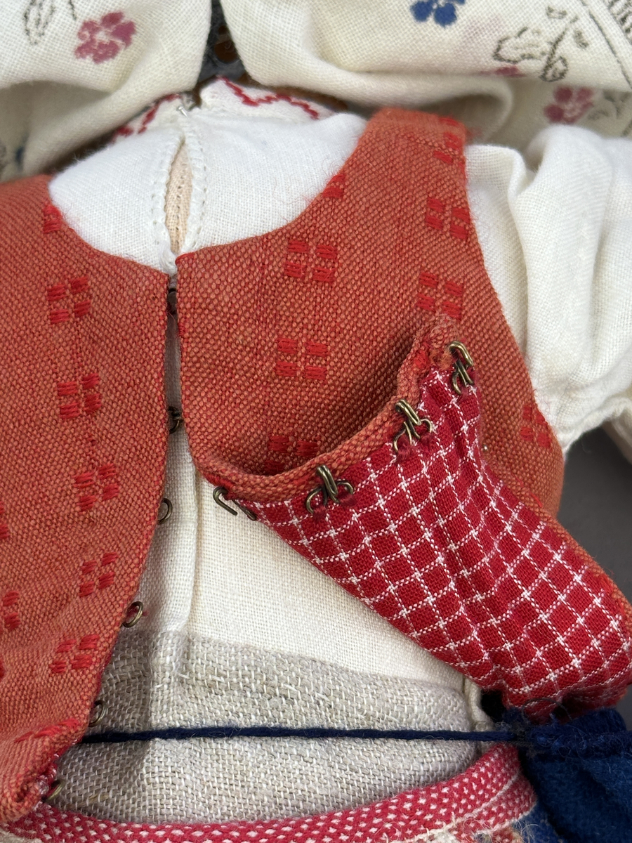 Docka klädd i kvinnlig sockendräkt från Östervallskogs socken i Värmland. Kroppen är handsydd i linnetyg och stoppad med textilt material. Huvud och händer är av lergods. Kläder handsydda av handvävda tyger förutom skjorta, underkjol och halskläde, som är maskinvävt. Skjorta är broderad med myrgång runt halssprund, plattsöm på hals- och handlinning samt en blå stjälksöm i kanten på handlinningen. Halsklädet, med tryck i gult, rosa och beige, hålls ihop av en sölja i stål med punsat mönster. Tyget till kjolen har oblekt linvarp med ullinslag i blått, grönt, rött och oblekt. Kjol sydd med lagda veck runtom, utom under förkläde, vars linning består av ett bomullsband. Förkläde, vävt med bomullsvarp och ullinslag i rött och vitt, har också lagda veck vid midja och bomullsband i rips till knytband. I midjan sitter en väska i blå och röd vadmal. Livstycket, vävt med bomullsvarp och lininslag, är vändsytt/fodrat med ett rutigt bomullstyg och knäpps med 5 hyskar & hakar. Ullstrumporna är handstickade i slätstickning med en kant upptill i 2-2 resår. Huvudet är prytt med en vit linnehätta kantad med en knypplad linnespets. Dockans fötter bär svarta skinnskor.

