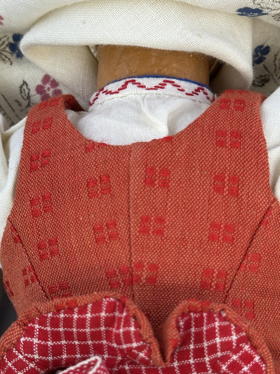 Docka klädd i kvinnlig sockendräkt från Östervallskogs socken i Värmland. Kroppen är handsydd i linnetyg och stoppad med textilt material. Huvud och händer är av lergods. Kläder handsydda av handvävda tyger förutom skjorta, underkjol och halskläde, som är maskinvävt. Skjorta är broderad med myrgång runt halssprund, plattsöm på hals- och handlinning samt en blå stjälksöm i kanten på handlinningen. Halsklädet, med tryck i gult, rosa och beige, hålls ihop av en sölja i stål med punsat mönster. Tyget till kjolen har oblekt linvarp med ullinslag i blått, grönt, rött och oblekt. Kjol sydd med lagda veck runtom, utom under förkläde, vars linning består av ett bomullsband. Förkläde, vävt med bomullsvarp och ullinslag i rött och vitt, har också lagda veck vid midja och bomullsband i rips till knytband. I midjan sitter en väska i blå och röd vadmal. Livstycket, vävt med bomullsvarp och lininslag, är vändsytt/fodrat med ett rutigt bomullstyg och knäpps med 5 hyskar & hakar. Ullstrumporna är handstickade i slätstickning med en kant upptill i 2-2 resår. Huvudet är prytt med en vit linnehätta kantad med en knypplad linnespets. Dockans fötter bär svarta skinnskor.

