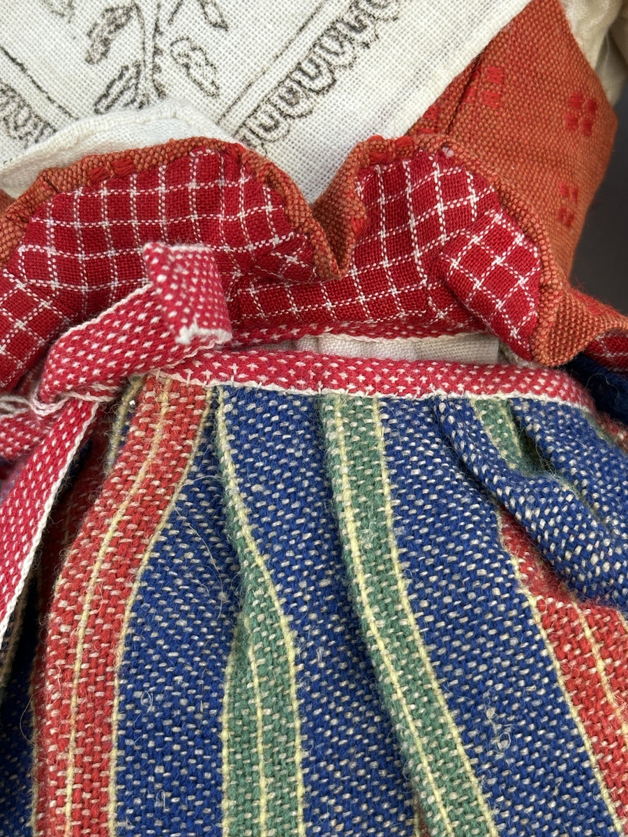 Docka klädd i kvinnlig sockendräkt från Östervallskogs socken i Värmland. Kroppen är handsydd i linnetyg och stoppad med textilt material. Huvud och händer är av lergods. Kläder handsydda av handvävda tyger förutom skjorta, underkjol och halskläde, som är maskinvävt. Skjorta är broderad med myrgång runt halssprund, plattsöm på hals- och handlinning samt en blå stjälksöm i kanten på handlinningen. Halsklädet, med tryck i gult, rosa och beige, hålls ihop av en sölja i stål med punsat mönster. Tyget till kjolen har oblekt linvarp med ullinslag i blått, grönt, rött och oblekt. Kjol sydd med lagda veck runtom, utom under förkläde, vars linning består av ett bomullsband. Förkläde, vävt med bomullsvarp och ullinslag i rött och vitt, har också lagda veck vid midja och bomullsband i rips till knytband. I midjan sitter en väska i blå och röd vadmal. Livstycket, vävt med bomullsvarp och lininslag, är vändsytt/fodrat med ett rutigt bomullstyg och knäpps med 5 hyskar & hakar. Ullstrumporna är handstickade i slätstickning med en kant upptill i 2-2 resår. Huvudet är prytt med en vit linnehätta kantad med en knypplad linnespets. Dockans fötter bär svarta skinnskor.

