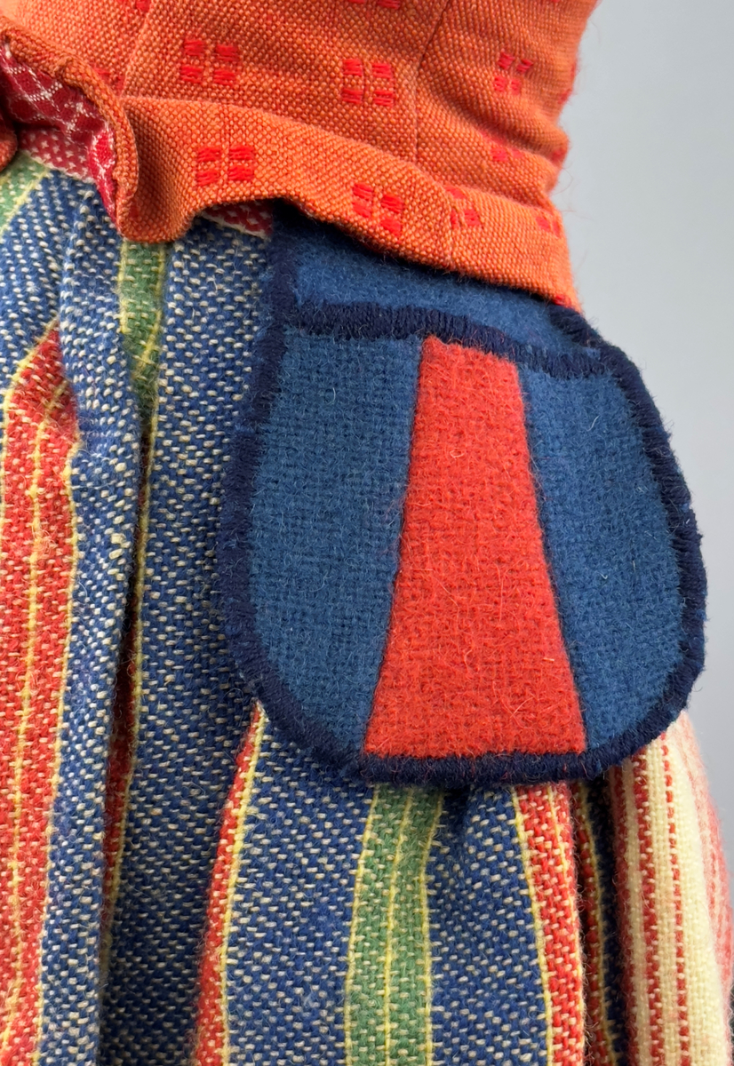 Docka klädd i kvinnlig sockendräkt från Östervallskogs socken i Värmland. Kroppen är handsydd i linnetyg och stoppad med textilt material. Huvud och händer är av lergods. Kläder handsydda av handvävda tyger förutom skjorta, underkjol och halskläde, som är maskinvävt. Skjorta är broderad med myrgång runt halssprund, plattsöm på hals- och handlinning samt en blå stjälksöm i kanten på handlinningen. Halsklädet, med tryck i gult, rosa och beige, hålls ihop av en sölja i stål med punsat mönster. Tyget till kjolen har oblekt linvarp med ullinslag i blått, grönt, rött och oblekt. Kjol sydd med lagda veck runtom, utom under förkläde, vars linning består av ett bomullsband. Förkläde, vävt med bomullsvarp och ullinslag i rött och vitt, har också lagda veck vid midja och bomullsband i rips till knytband. I midjan sitter en väska i blå och röd vadmal. Livstycket, vävt med bomullsvarp och lininslag, är vändsytt/fodrat med ett rutigt bomullstyg och knäpps med 5 hyskar & hakar. Ullstrumporna är handstickade i slätstickning med en kant upptill i 2-2 resår. Huvudet är prytt med en vit linnehätta kantad med en knypplad linnespets. Dockans fötter bär svarta skinnskor.

