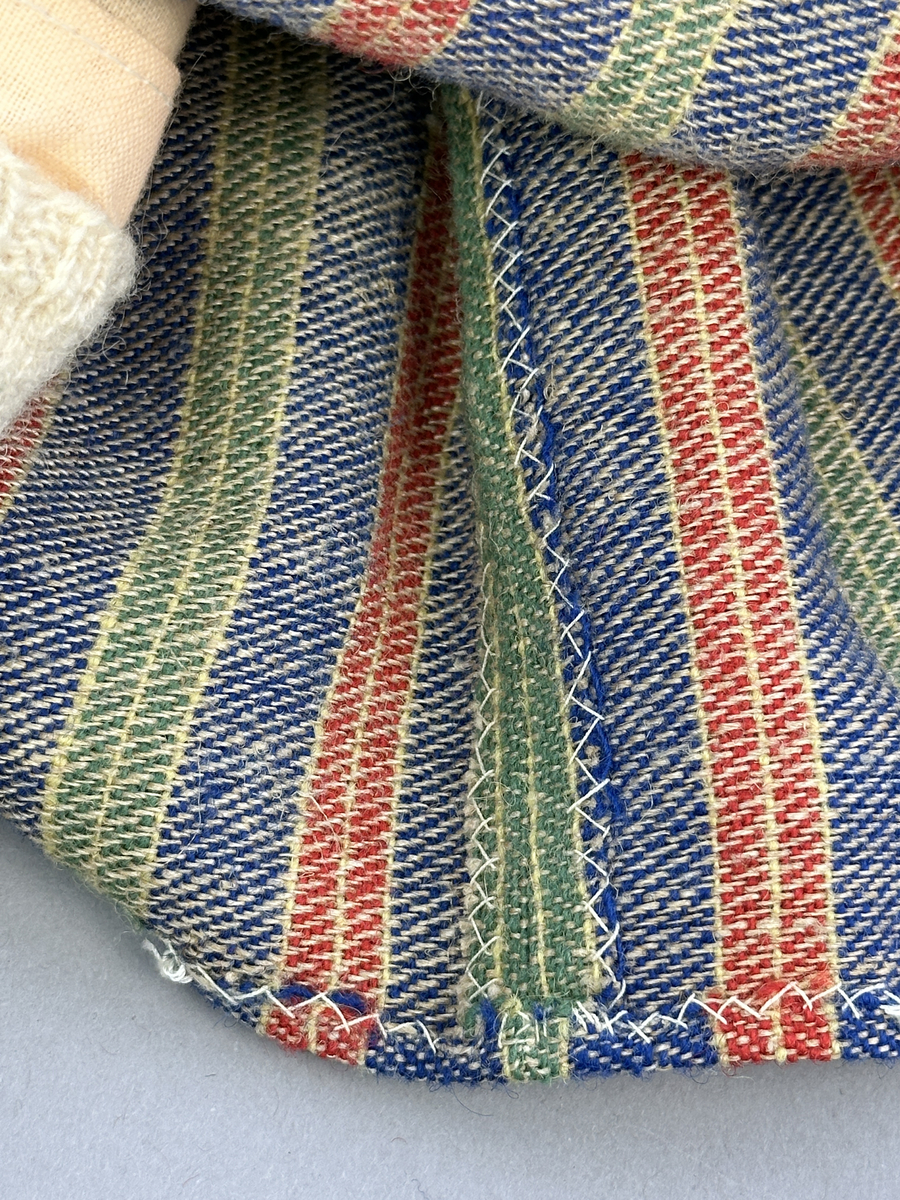 Docka klädd i kvinnlig sockendräkt från Östervallskogs socken i Värmland. Kroppen är handsydd i linnetyg och stoppad med textilt material. Huvud och händer är av lergods. Kläder handsydda av handvävda tyger förutom skjorta, underkjol och halskläde, som är maskinvävt. Skjorta är broderad med myrgång runt halssprund, plattsöm på hals- och handlinning samt en blå stjälksöm i kanten på handlinningen. Halsklädet, med tryck i gult, rosa och beige, hålls ihop av en sölja i stål med punsat mönster. Tyget till kjolen har oblekt linvarp med ullinslag i blått, grönt, rött och oblekt. Kjol sydd med lagda veck runtom, utom under förkläde, vars linning består av ett bomullsband. Förkläde, vävt med bomullsvarp och ullinslag i rött och vitt, har också lagda veck vid midja och bomullsband i rips till knytband. I midjan sitter en väska i blå och röd vadmal. Livstycket, vävt med bomullsvarp och lininslag, är vändsytt/fodrat med ett rutigt bomullstyg och knäpps med 5 hyskar & hakar. Ullstrumporna är handstickade i slätstickning med en kant upptill i 2-2 resår. Huvudet är prytt med en vit linnehätta kantad med en knypplad linnespets. Dockans fötter bär svarta skinnskor.

