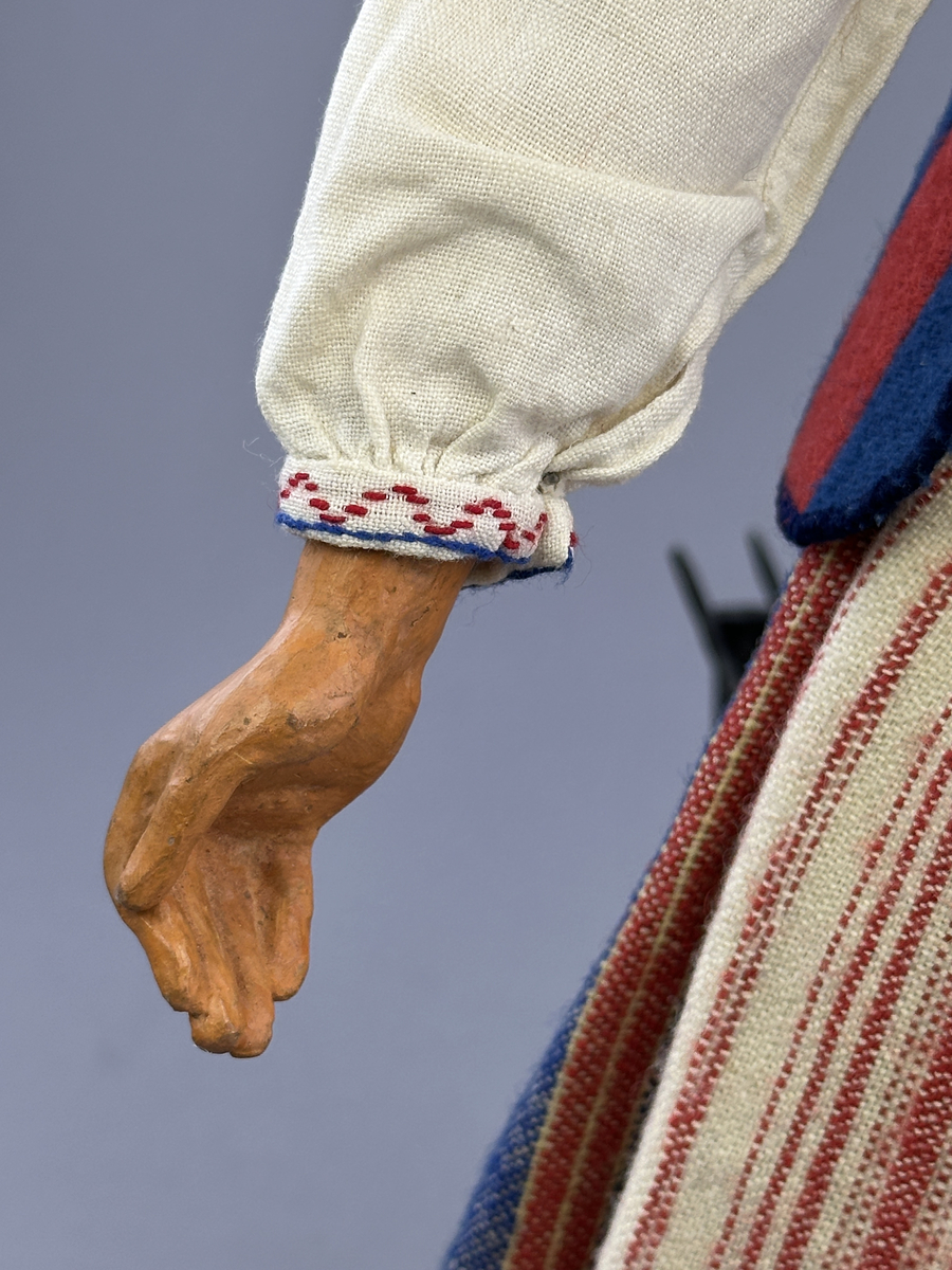 Docka klädd i kvinnlig sockendräkt från Östervallskogs socken i Värmland. Kroppen är handsydd i linnetyg och stoppad med textilt material. Huvud och händer är av lergods. Kläder handsydda av handvävda tyger förutom skjorta, underkjol och halskläde, som är maskinvävt. Skjorta är broderad med myrgång runt halssprund, plattsöm på hals- och handlinning samt en blå stjälksöm i kanten på handlinningen. Halsklädet, med tryck i gult, rosa och beige, hålls ihop av en sölja i stål med punsat mönster. Tyget till kjolen har oblekt linvarp med ullinslag i blått, grönt, rött och oblekt. Kjol sydd med lagda veck runtom, utom under förkläde, vars linning består av ett bomullsband. Förkläde, vävt med bomullsvarp och ullinslag i rött och vitt, har också lagda veck vid midja och bomullsband i rips till knytband. I midjan sitter en väska i blå och röd vadmal. Livstycket, vävt med bomullsvarp och lininslag, är vändsytt/fodrat med ett rutigt bomullstyg och knäpps med 5 hyskar & hakar. Ullstrumporna är handstickade i slätstickning med en kant upptill i 2-2 resår. Huvudet är prytt med en vit linnehätta kantad med en knypplad linnespets. Dockans fötter bär svarta skinnskor.


