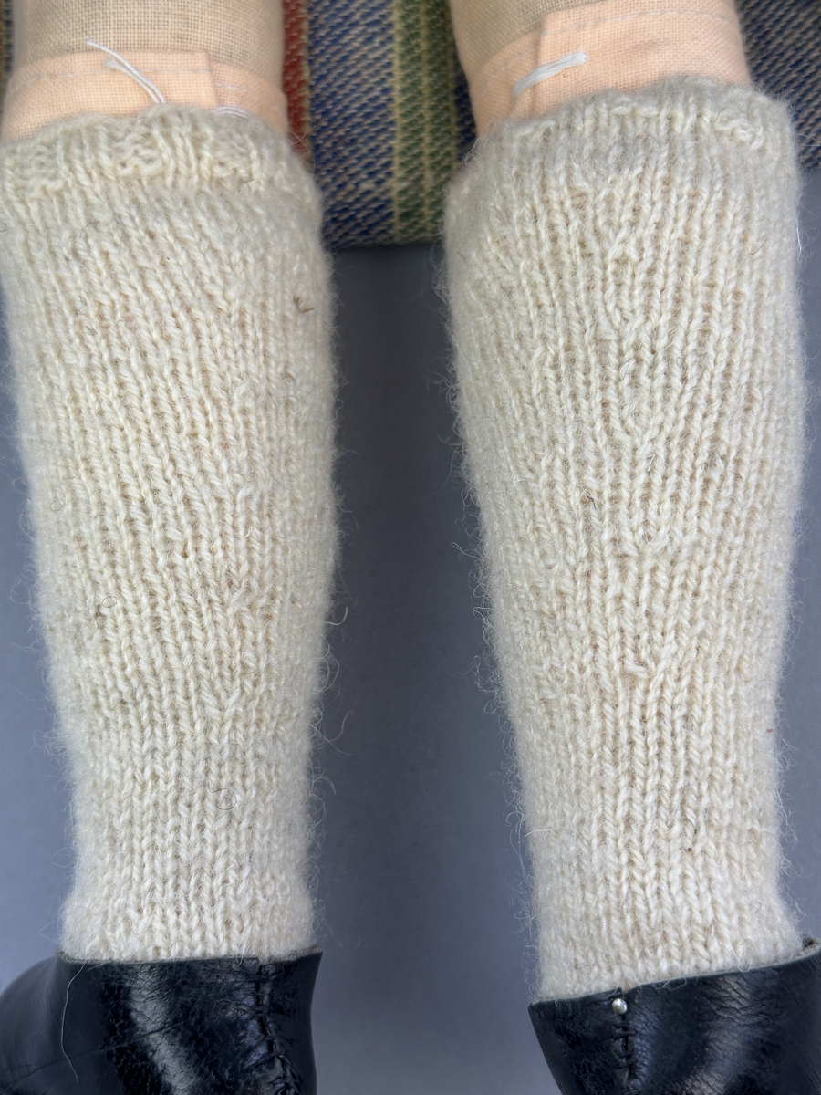 Docka klädd i kvinnlig sockendräkt från Östervallskogs socken i Värmland. Kroppen är handsydd i linnetyg och stoppad med textilt material. Huvud och händer är av lergods. Kläder handsydda av handvävda tyger förutom skjorta, underkjol och halskläde, som är maskinvävt. Skjorta är broderad med myrgång runt halssprund, plattsöm på hals- och handlinning samt en blå stjälksöm i kanten på handlinningen. Halsklädet, med tryck i gult, rosa och beige, hålls ihop av en sölja i stål med punsat mönster. Tyget till kjolen har oblekt linvarp med ullinslag i blått, grönt, rött och oblekt. Kjol sydd med lagda veck runtom, utom under förkläde, vars linning består av ett bomullsband. Förkläde, vävt med bomullsvarp och ullinslag i rött och vitt, har också lagda veck vid midja och bomullsband i rips till knytband. I midjan sitter en väska i blå och röd vadmal. Livstycket, vävt med bomullsvarp och lininslag, är vändsytt/fodrat med ett rutigt bomullstyg och knäpps med 5 hyskar & hakar. Ullstrumporna är handstickade i slätstickning med en kant upptill i 2-2 resår. Huvudet är prytt med en vit linnehätta kantad med en knypplad linnespets. Dockans fötter bär svarta skinnskor.

