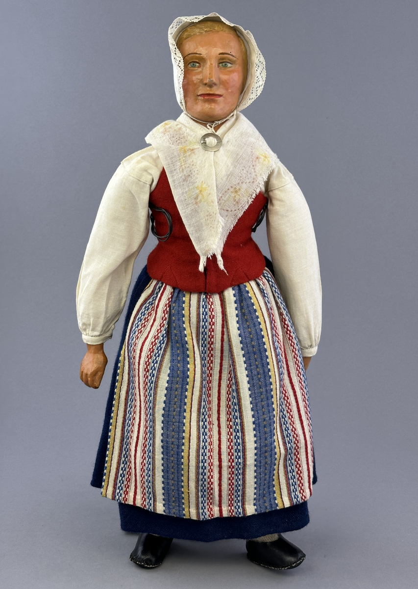Docka klädd i kvinnlig sockendräkt från Ulleruds socken i Värmland. Kroppen är handsydd i linnetyg och stoppad med textilt material. Huvud och händer är av lergods. Kläder handsydda av handvävda tyger förutom skjorta, underkjol och halskläde, som är maskinvävt. Skjorta, broderad med myrgång runt sprund, har halsspjäll, halslinning och handlinning. Halsklädet, med tryck i gult, rosa och beige, hålls ihop av en sölja i nysilver med graverat mönster. Kjol sydd med lagda veck runtom, utom under förkläde, vars linning består av ett bomullsband. Förkläde, vävt i tuskaft och munkabälte, har lagda veck vid midja och bomullsband i rips till knytband. Livstycket är vändsytt/fodrat med ett rutigt bomullstyg och knäpps med 6 hyskar & hakar. Ullstrumporna är handstickade i slätstickning med en kant upptill i 2-2 resår. Huvudet är prytt med en vit linnehätta kantad med en knypplad linnespets. Dockans fötter bär svarta skinnskor.