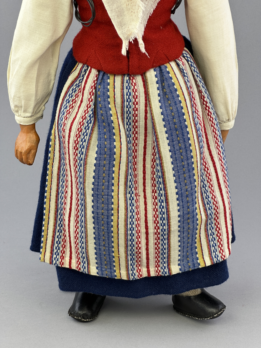 Docka klädd i kvinnlig sockendräkt från Ulleruds socken i Värmland. Kroppen är handsydd i linnetyg och stoppad med textilt material. Huvud och händer är av lergods. Kläder handsydda av handvävda tyger förutom skjorta, underkjol och halskläde, som är maskinvävt. Skjorta, broderad med myrgång runt sprund, har halsspjäll, halslinning och handlinning. Halsklädet, med tryck i gult, rosa och beige, hålls ihop av en sölja i nysilver med graverat mönster. Kjol sydd med lagda veck runtom, utom under förkläde, vars linning består av ett bomullsband. Förkläde, vävt i tuskaft och munkabälte, har lagda veck vid midja och bomullsband i rips till knytband. Livstycket är vändsytt/fodrat med ett rutigt bomullstyg och knäpps med 6 hyskar & hakar. Ullstrumporna är handstickade i slätstickning med en kant upptill i 2-2 resår. Huvudet är prytt med en vit linnehätta kantad med en knypplad linnespets. Dockans fötter bär svarta skinnskor.