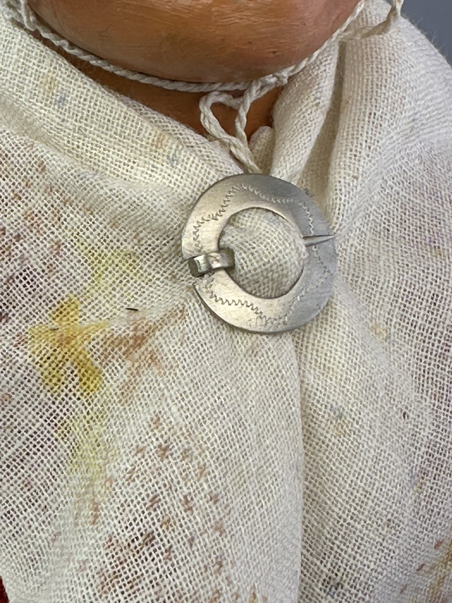 Docka klädd i kvinnlig sockendräkt från Ulleruds socken i Värmland. Kroppen är handsydd i linnetyg och stoppad med textilt material. Huvud och händer är av lergods. Kläder handsydda av handvävda tyger förutom skjorta, underkjol och halskläde, som är maskinvävt. Skjorta, broderad med myrgång runt sprund, har halsspjäll, halslinning och handlinning. Halsklädet, med tryck i gult, rosa och beige, hålls ihop av en sölja i nysilver med graverat mönster. Kjol sydd med lagda veck runtom, utom under förkläde, vars linning består av ett bomullsband. Förkläde, vävt i tuskaft och munkabälte, har lagda veck vid midja och bomullsband i rips till knytband. Livstycket är vändsytt/fodrat med ett rutigt bomullstyg och knäpps med 6 hyskar & hakar. Ullstrumporna är handstickade i slätstickning med en kant upptill i 2-2 resår. Huvudet är prytt med en vit linnehätta kantad med en knypplad linnespets. Dockans fötter bär svarta skinnskor.
