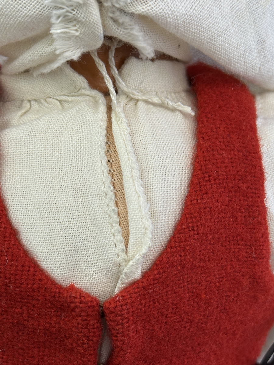 Docka klädd i kvinnlig sockendräkt från Ulleruds socken i Värmland. Kroppen är handsydd i linnetyg och stoppad med textilt material. Huvud och händer är av lergods. Kläder handsydda av handvävda tyger förutom skjorta, underkjol och halskläde, som är maskinvävt. Skjorta, broderad med myrgång runt sprund, har halsspjäll, halslinning och handlinning. Halsklädet, med tryck i gult, rosa och beige, hålls ihop av en sölja i nysilver med graverat mönster. Kjol sydd med lagda veck runtom, utom under förkläde, vars linning består av ett bomullsband. Förkläde, vävt i tuskaft och munkabälte, har lagda veck vid midja och bomullsband i rips till knytband. Livstycket är vändsytt/fodrat med ett rutigt bomullstyg och knäpps med 6 hyskar & hakar. Ullstrumporna är handstickade i slätstickning med en kant upptill i 2-2 resår. Huvudet är prytt med en vit linnehätta kantad med en knypplad linnespets. Dockans fötter bär svarta skinnskor.