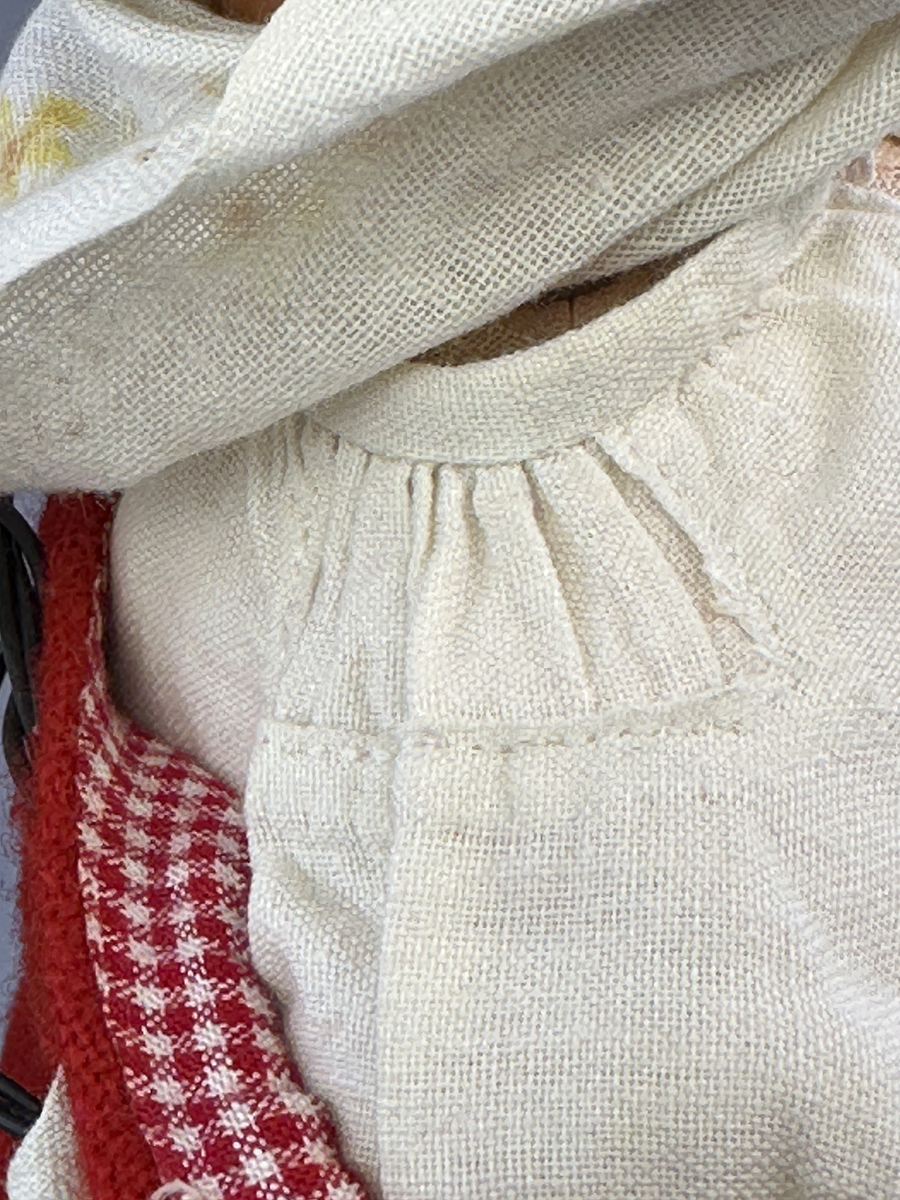 Docka klädd i kvinnlig sockendräkt från Ulleruds socken i Värmland. Kroppen är handsydd i linnetyg och stoppad med textilt material. Huvud och händer är av lergods. Kläder handsydda av handvävda tyger förutom skjorta, underkjol och halskläde, som är maskinvävt. Skjorta, broderad med myrgång runt sprund, har halsspjäll, halslinning och handlinning. Halsklädet, med tryck i gult, rosa och beige, hålls ihop av en sölja i nysilver med graverat mönster. Kjol sydd med lagda veck runtom, utom under förkläde, vars linning består av ett bomullsband. Förkläde, vävt i tuskaft och munkabälte, har lagda veck vid midja och bomullsband i rips till knytband. Livstycket är vändsytt/fodrat med ett rutigt bomullstyg och knäpps med 6 hyskar & hakar. Ullstrumporna är handstickade i slätstickning med en kant upptill i 2-2 resår. Huvudet är prytt med en vit linnehätta kantad med en knypplad linnespets. Dockans fötter bär svarta skinnskor.